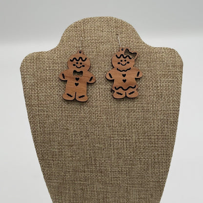 Two wooden gingerbread man earrings are displayed on a textured beige stand. The left earring has a bowtie and buttons, while the right earring features a bow on its head and a heart on the chest. Both are decorated with black accents, giving them a festive, playful appearance.