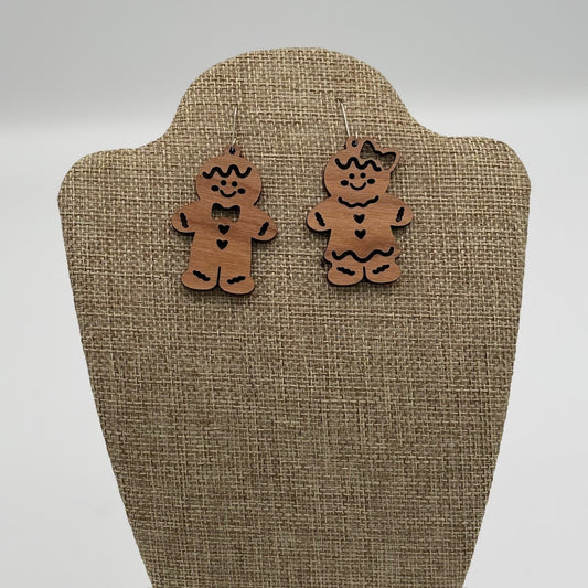 Two wooden gingerbread man earrings are displayed on a textured beige stand. The left earring has a bowtie and buttons, while the right earring features a bow on its head and a heart on the chest. Both are decorated with black accents, giving them a festive, playful appearance.
