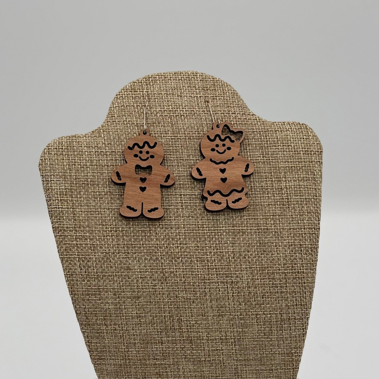 Close-up of wooden gingerbread man and woman earrings displayed on a beige textured stand. The man has a bowtie and buttons, while the woman has a bow and heart details. Both figures feature black accents and a festive design. The background is a plain, light gray.