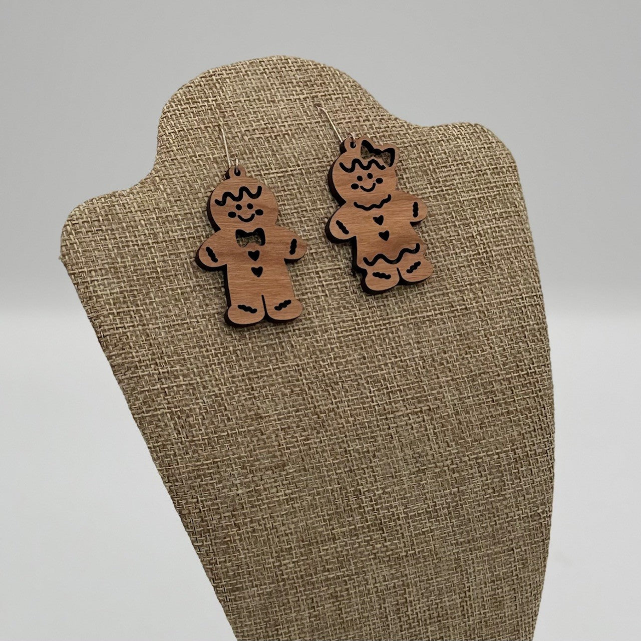 A pair of wooden gingerbread man earrings are displayed on a beige fabric-covered stand. Each earring features a smiling face, black bow tie, buttons, and wavy icing details. The background is plain and gray, highlighting the earrings playful and festive design.