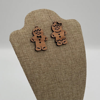 A pair of wooden gingerbread man earrings are displayed on a beige fabric-covered stand. Each earring features a smiling face, black bow tie, buttons, and wavy icing details. The background is plain and gray, highlighting the earrings playful and festive design.