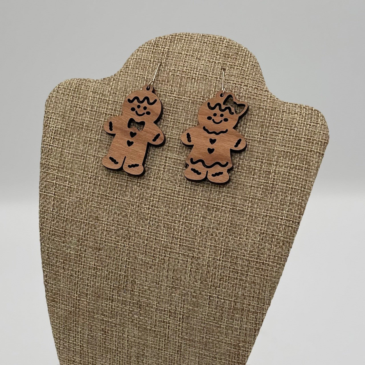A pair of gingerbread man and woman wooden earrings with engraved details, including eyes, buttons, and bows, are displayed on a beige textured stand. The background is plain and light gray.