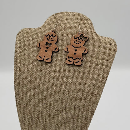 A pair of gingerbread man and woman wooden earrings with engraved details, including eyes, buttons, and bows, are displayed on a beige textured stand. The background is plain and light gray.