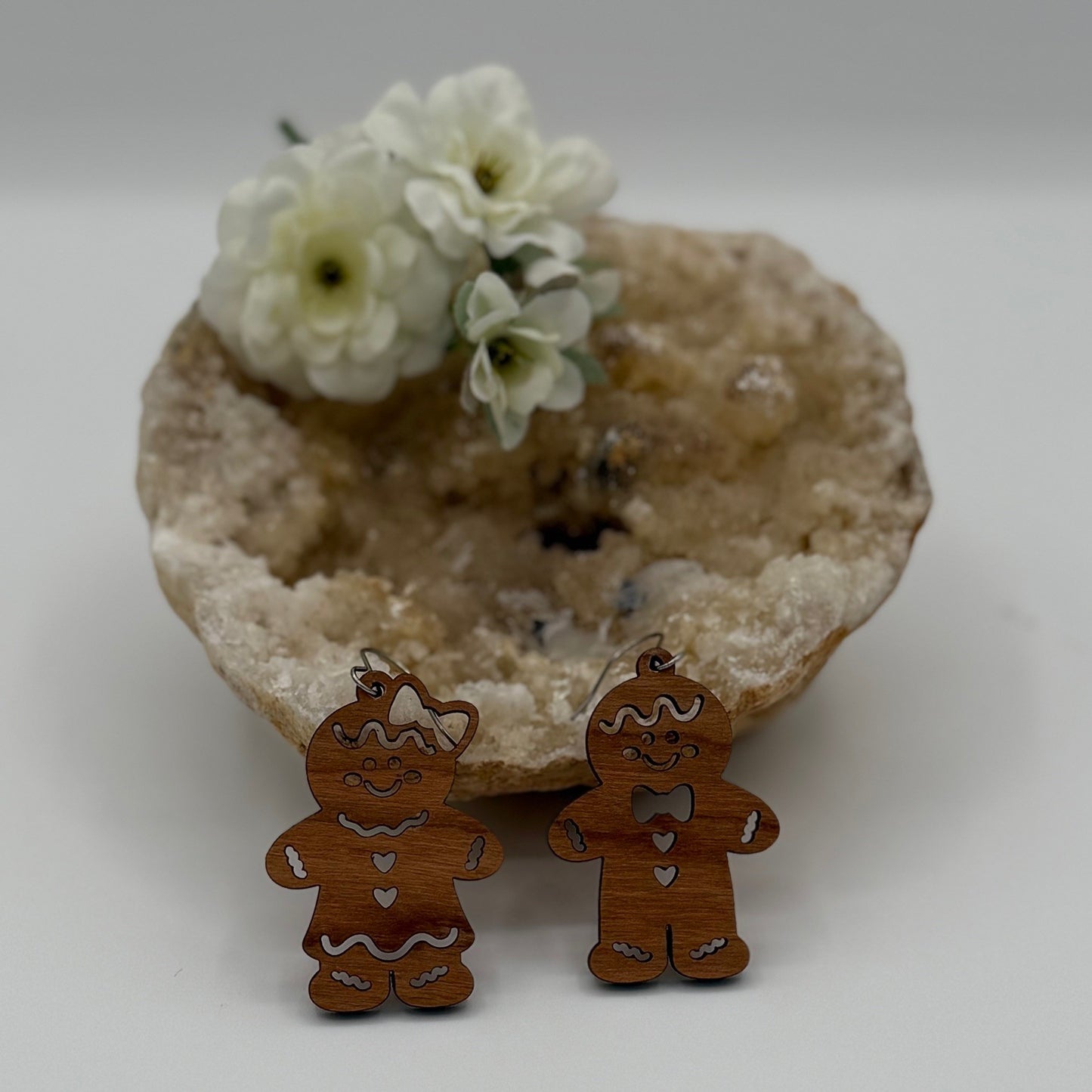 Two wooden gingerbread ornaments, one with a bow and the other with a bow tie, stand in front of a geode. Behind them, white flowers rest on the geodes surface, adding a delicate touch to the scene.