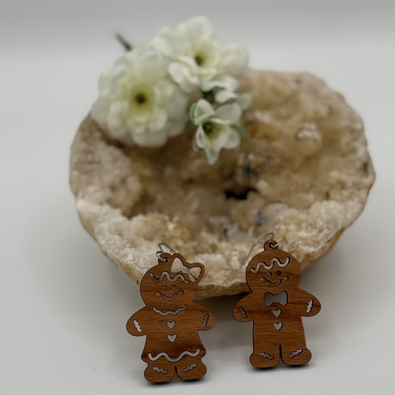 Two wooden gingerbread man ornaments with white icing details stand in front of a rough, circular stone bowl. White flowers are placed inside the bowl, adding a decorative touch to the scene. The background is softly blurred, highlighting the ornaments and flowers in the foreground.