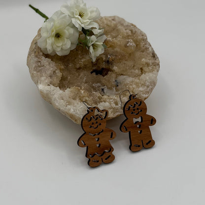 Two wooden gingerbread man earrings are placed in front of a textured, light-colored rock. The rock has a small cluster of white flowers resting on top. The gingerbread figures have distinct expressions, one with a bow tie and the other with a scarf.