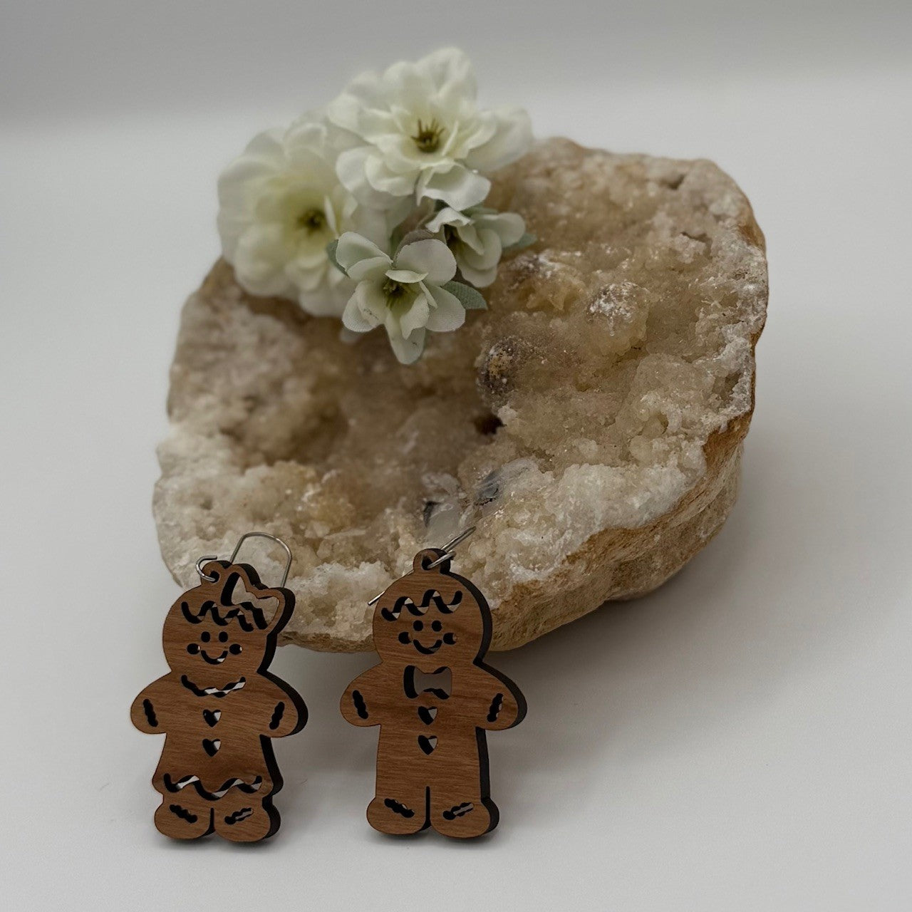 Wooden gingerbread man and woman earrings are displayed in front of a large, textured crystal rock. White flowers are placed on top of the rock, adding a delicate touch to the composition. The earrings have engraved details and attached hooks for wearing.