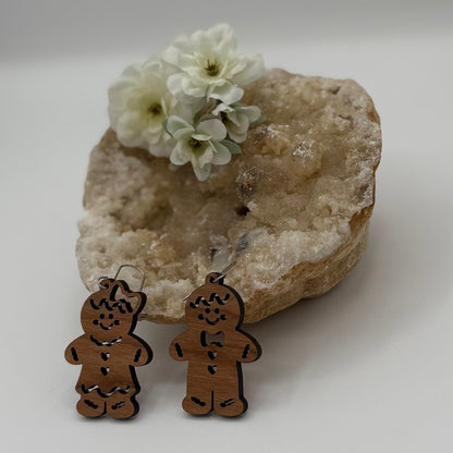 Wooden gingerbread man and woman earrings are displayed in front of a large, textured crystal rock. White flowers are placed on top of the rock, adding a delicate touch to the composition. The earrings have engraved details and attached hooks for wearing.