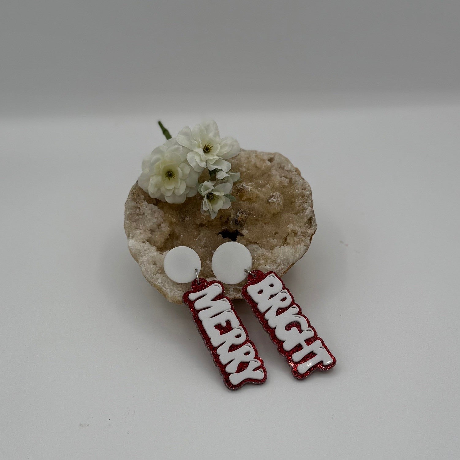 A decorative display featuring white flowers placed in a textured bowl. Adjacent to the flowers are two circular, white earrings with red-glittered text: one reads MERRY, and the other reads BRIGHT. The background is a soft, neutral color.