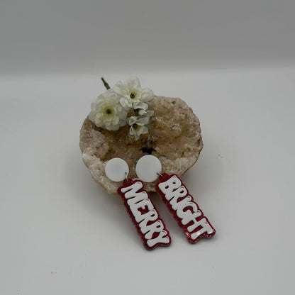 A decorative display featuring white flowers placed in a textured bowl. Adjacent to the flowers are two circular, white earrings with red-glittered text: one reads MERRY, and the other reads BRIGHT. The background is a soft, neutral color.