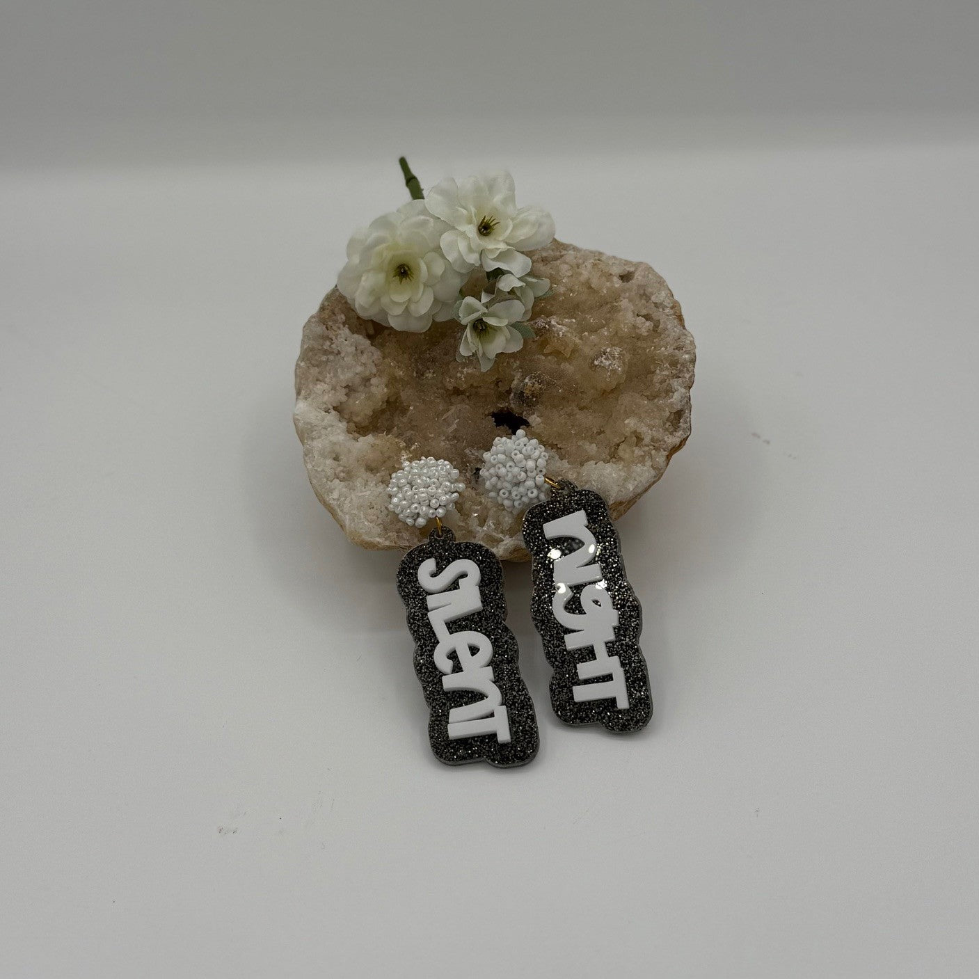 A decorative stone with small white flowers holds a pair of black earrings featuring the words silent night in white. The earrings hang from spherical, pearl-like studs, creating a festive and elegant display against a simple gray background.