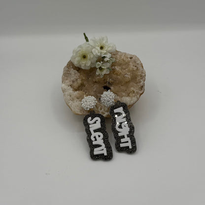 A decorative stone with small white flowers holds a pair of black earrings featuring the words silent night in white. The earrings hang from spherical, pearl-like studs, creating a festive and elegant display against a simple gray background.