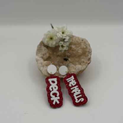 A pair of red glittery earrings with white symbols rests on a beige, textured stone-like surface. Behind them, small white flowers are positioned above the stone, adding a natural decorative element. The background is a neutral light gray, creating a simple and clean display.