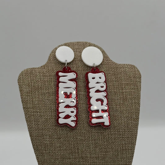 The image shows a pair of festive earrings on a display stand. The earrings are shaped like vertical rectangles with the words MERRY and BRIGHT in white letters on a red glittery background. The top part of each earring is a round, white stud.