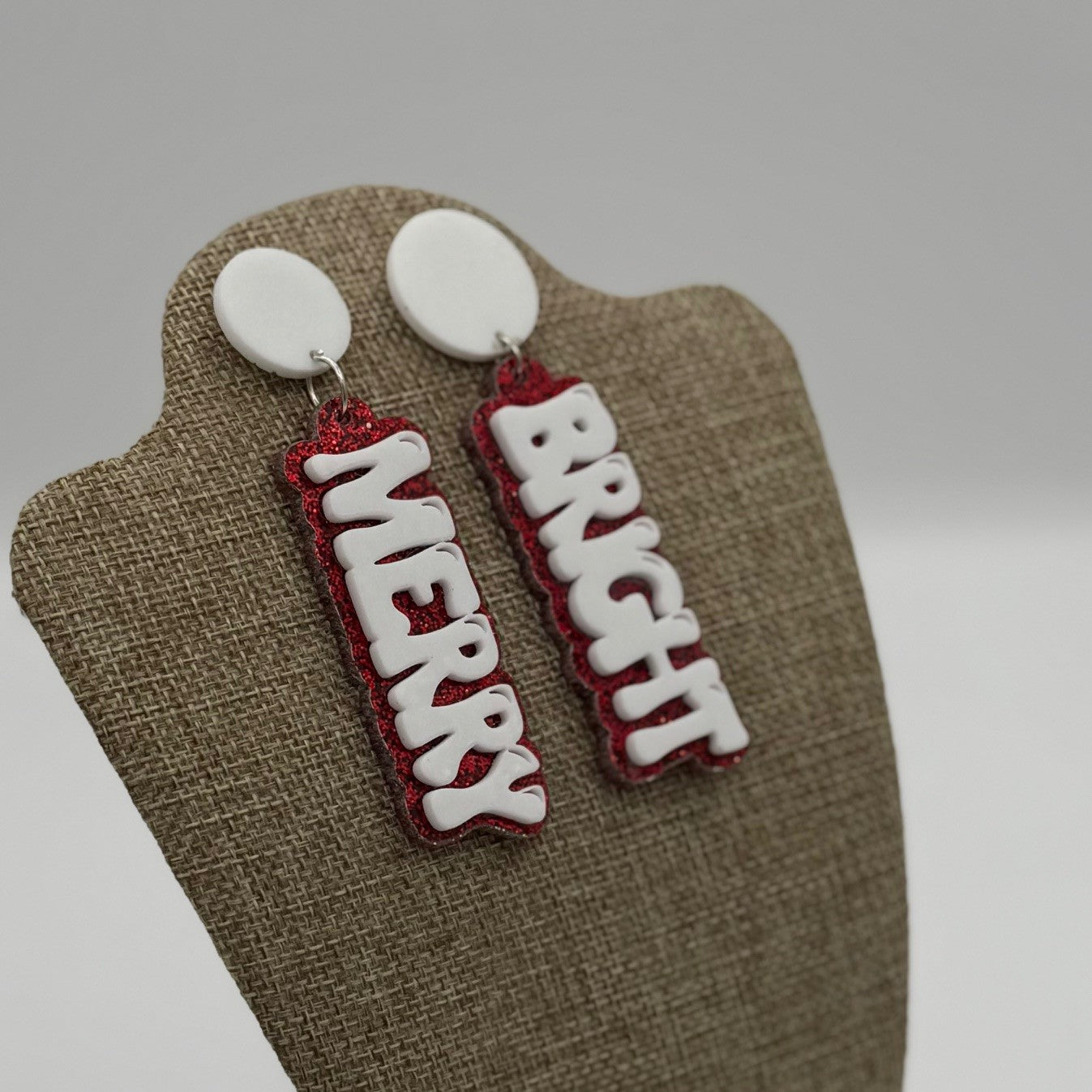A pair of earrings displayed on a textured stand. Each earring has a circular white stud and a dangling rectangular charm. One charm reads MERRY and the other BRIGHT in bold white letters with red sparkly borders. The background is plain and neutral.