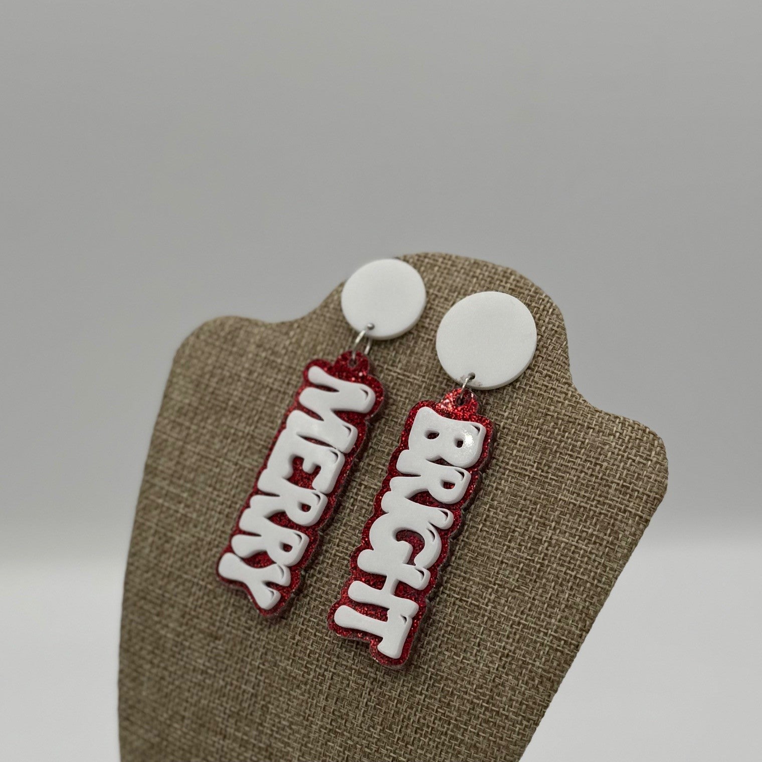 Two festive earrings hang on a display stand. Each earring has a white circular top with a dangling word: MERRY on the left and BRIGHT on the right. The words are in white letters outlined in red glittery material, giving a cheerful holiday vibe. The background is a soft gray.