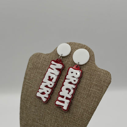 Two festive earrings hang on a display stand. Each earring has a white circular top with a dangling word: MERRY on the left and BRIGHT on the right. The words are in white letters outlined in red glittery material, giving a cheerful holiday vibe. The background is a soft gray.