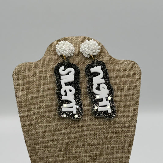 A pair of earrings displayed on a stand. Each earring has a round, white, beaded stud with a dangling pendant. The pendants are black with glitter and have the words Silent and Night in bold, white letters.