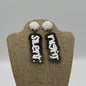 A pair of earrings displayed on a stand. Each earring has a round, white, beaded stud with a dangling pendant. The pendants are black with glitter and have the words Silent and Night in bold, white letters.