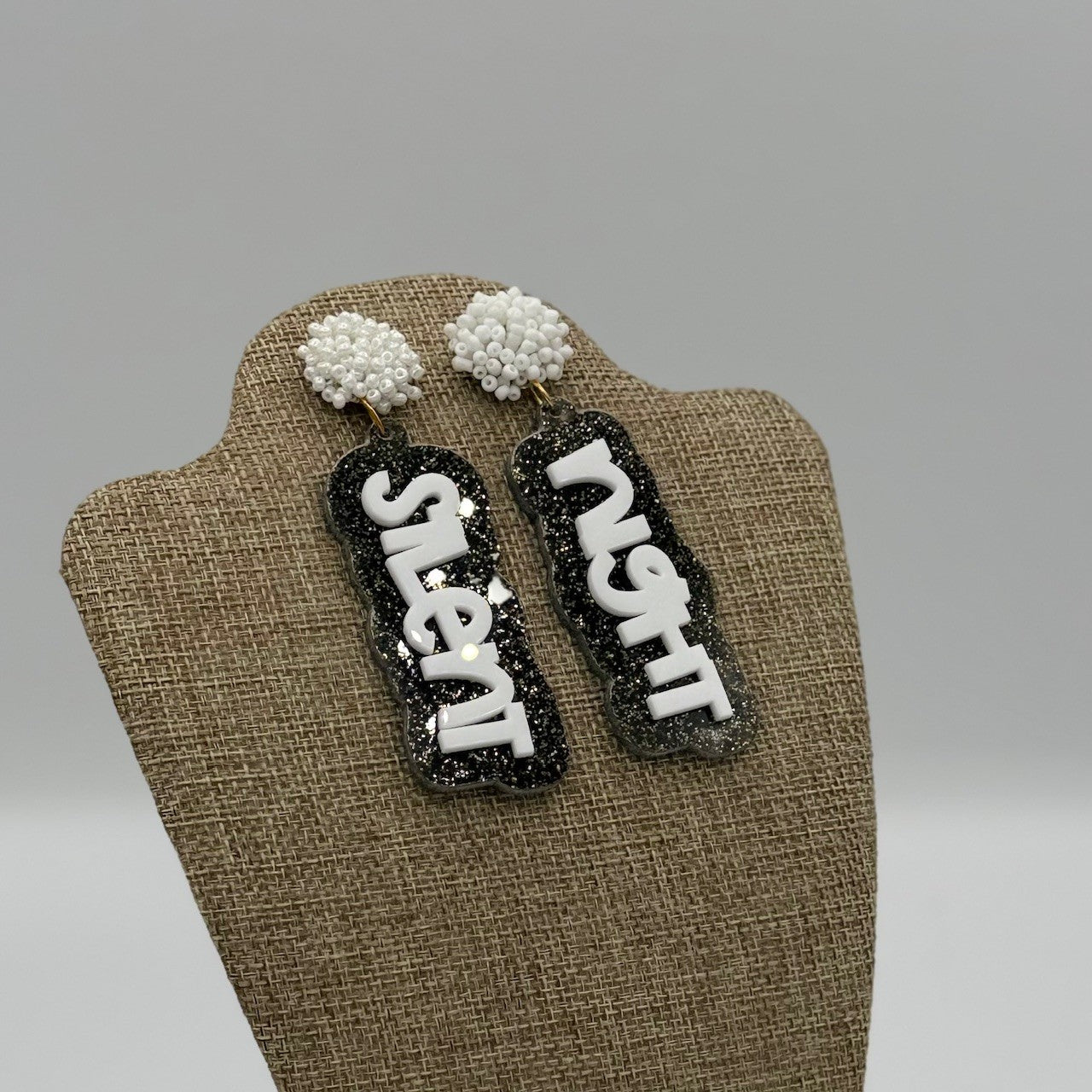 A display mannequin holds earrings with the words silent night. The earrings have black and white glittery backgrounds with the text in bold white letters. The top part of the earrings resembles small clusters of white beads. The background is plain gray.