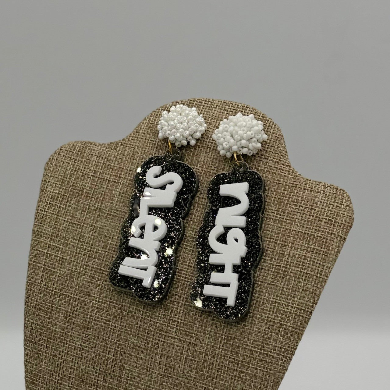 Earrings displayed on a beige stand. The top parts are clusters of small white beads. The dangling sections are glittery black with white bubble lettering spelling Silent Night vertically.