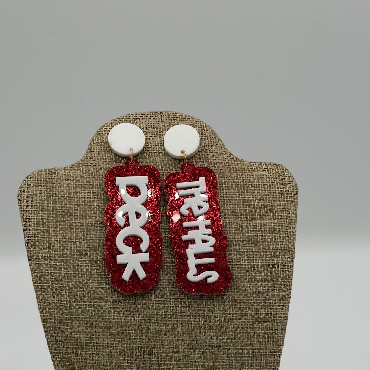 A pair of earrings on display, each with a white circular top and a dangling red glittery rectangular piece. The left earring reads DECK in white letters, and the right one reads THE HALLS, completing the festive phrase. The background is neutral, highlighting the earrings.