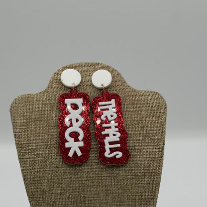 A pair of earrings on display, each with a white circular top and a dangling red glittery rectangular piece. The left earring reads DECK in white letters, and the right one reads THE HALLS, completing the festive phrase. The background is neutral, highlighting the earrings.