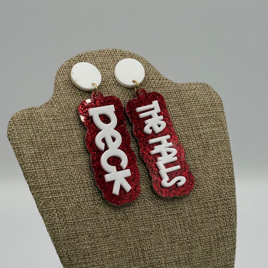 A pair of statement earrings with red glittery backgrounds. Each earring features white text; one reads Deck and the other The Halls. The earrings are displayed on a burlap-textured stand, against a neutral gray background.