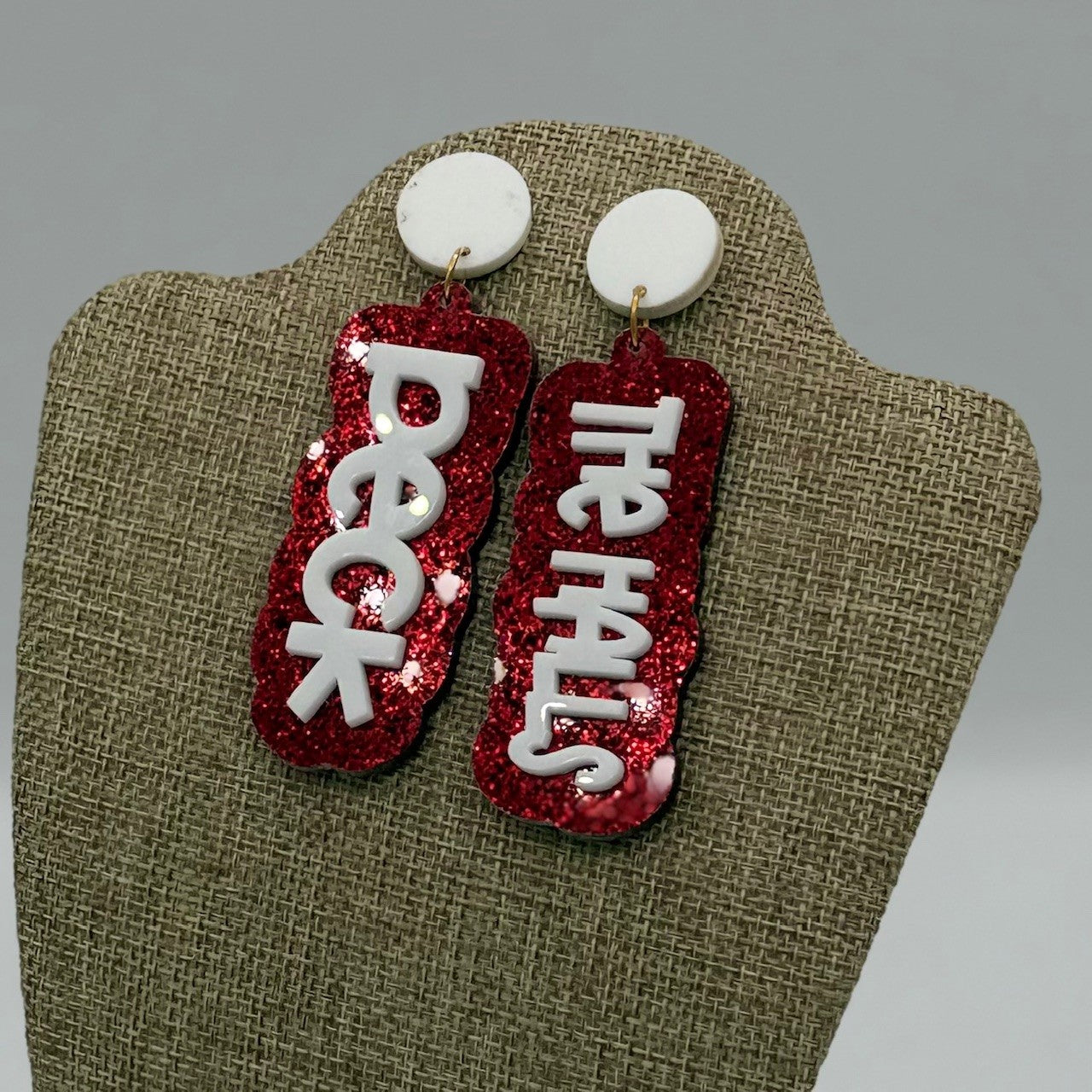 A pair of statement earrings with red glittery backgrounds. Each earring features white text; one reads Deck and the other The Halls. The earrings are displayed on a burlap-textured stand, against a neutral gray background.