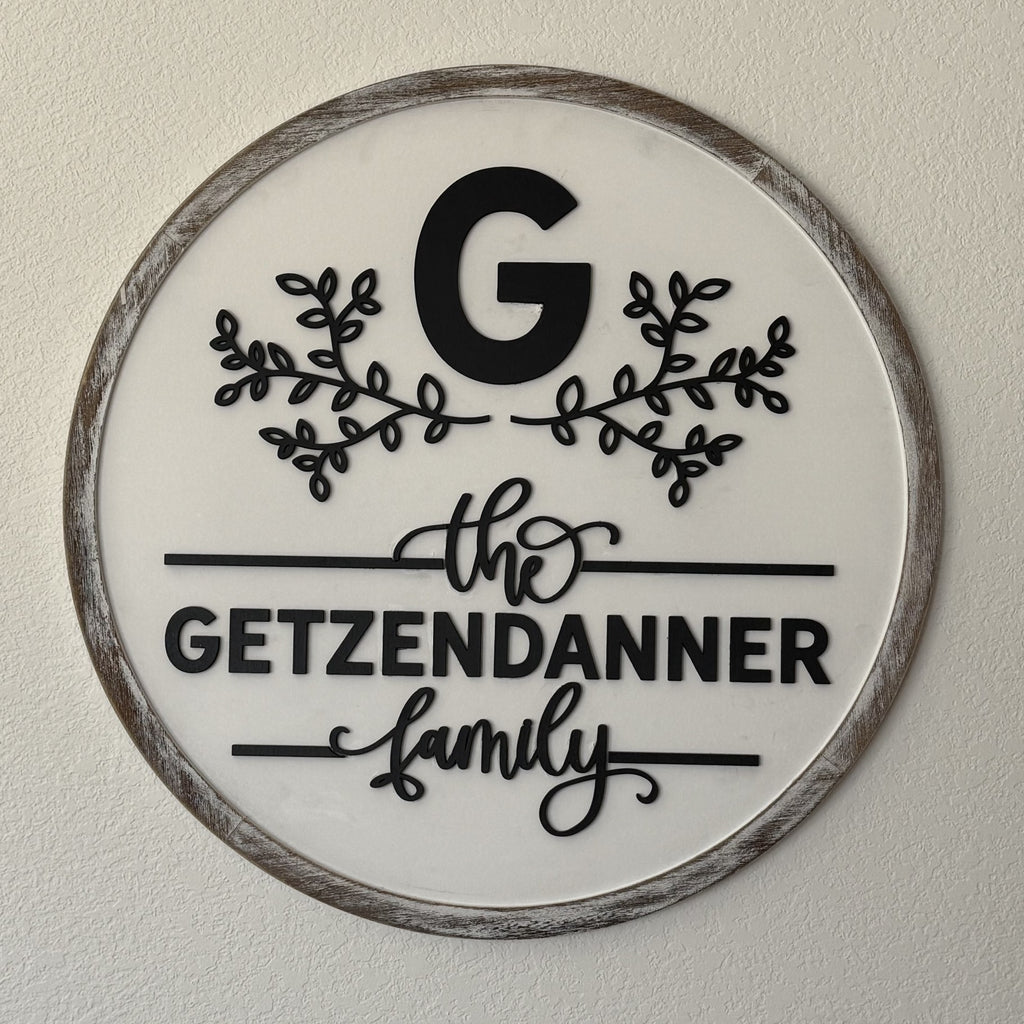 A round wall sign with a wooden frame features a large black letter G at the top, adorned with leafy branches on both sides. Below, in black script and bold type, the text reads the GETZENDANNER family against a white background.