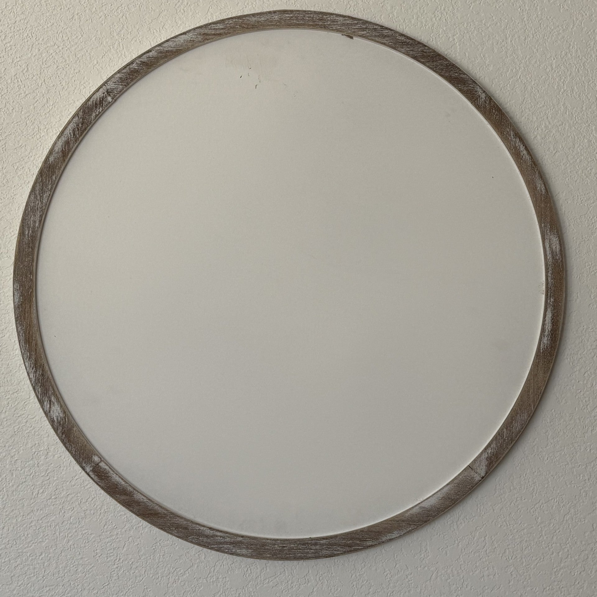 A round wall sign with a rustic wooden frame is mounted on a plain white wall. The frame has a weathered, distressed look, adding a touch of vintage charm. The sign is blank, showing the simplicity and minimalism of the decor.