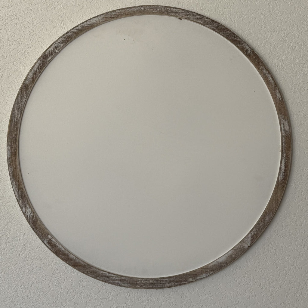 A round wall sign with a rustic wooden frame is mounted on a plain white wall. The frame has a weathered, distressed look, adding a touch of vintage charm. The sign is blank, showing the simplicity and minimalism of the decor.