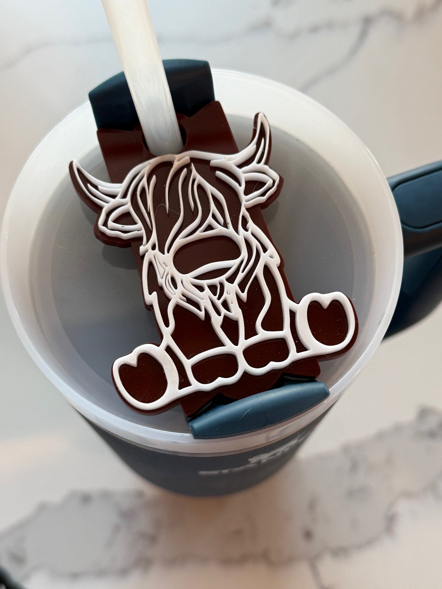 A milk jug topped with a latte features a chocolate decoration shaped like a highland cows face, outlined in white. The chocolate is placed on top of the drink with a straw emerging from the cup. The background shows a white, textured surface.