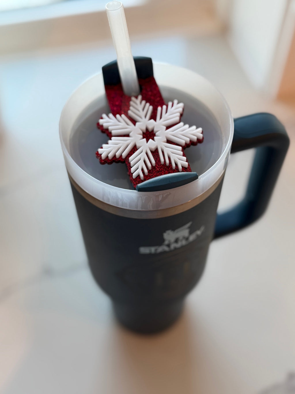 A large insulated mug with a handle is filled with a dark liquid. A straw is inserted into the mug. Resting on top is a red and white snowflake-shaped coaster. The mug is placed on a light-colored surface near a window, and it has a logo featuring a mountain and the word Stanley.