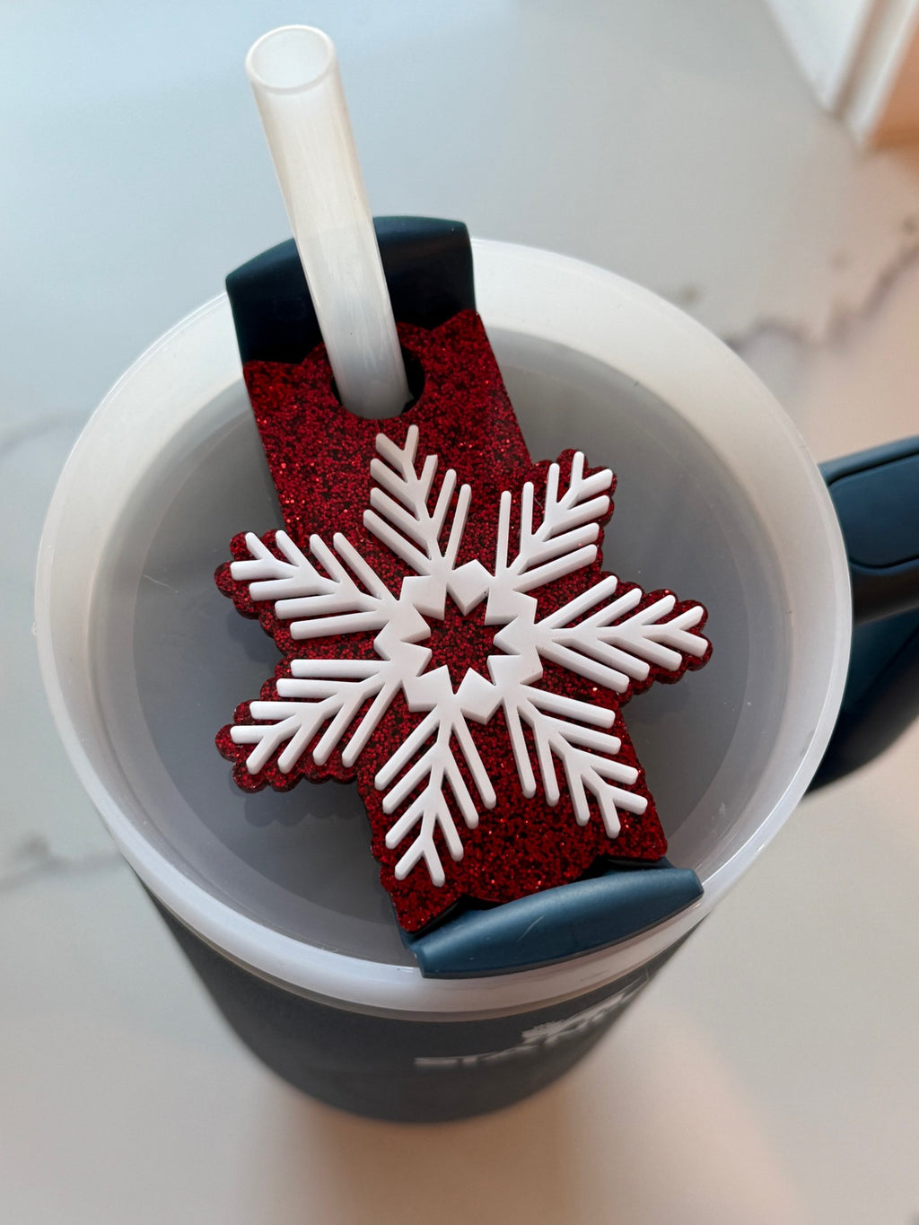 A close-up image of a dark cup with a lid and straw. On the lid, theres a red glittery decoration featuring a large white snowflake design. The background shows a blurred white and gray surface.