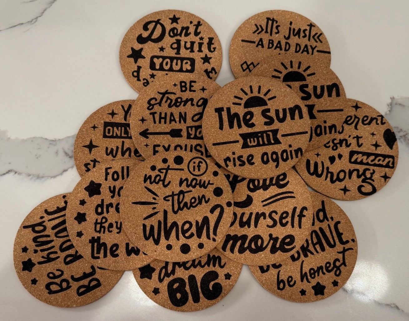 The Motivational Cork Coasters set includes four round pieces with inspirational phrases like Dont quit your daydream, Be stronger than your excuses, and Dream big in black lettering, adorned with stars and decorative elements to protect surfaces while inspiring you.