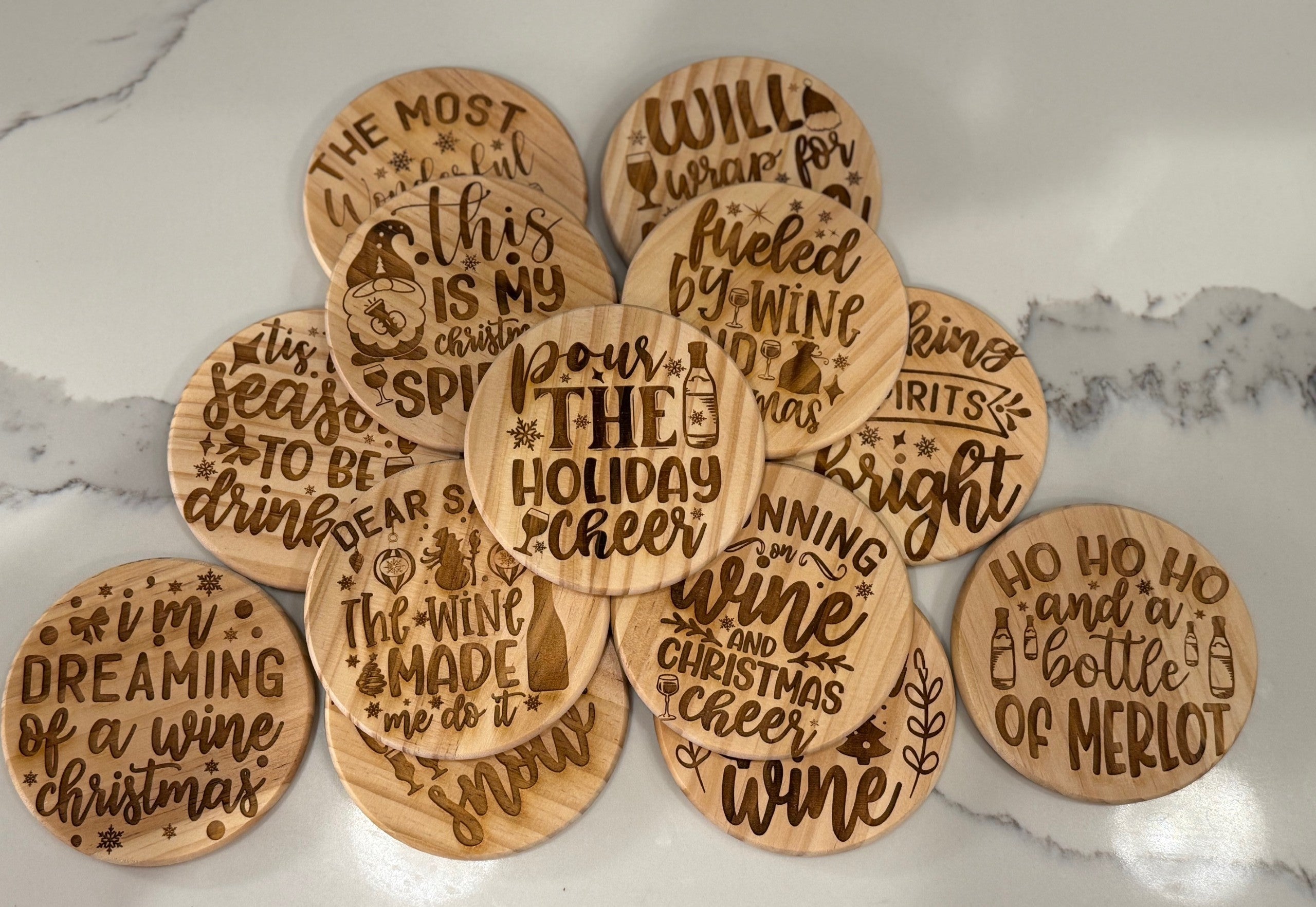 The Funny Wine Sayings set includes four round wooden coasters featuring humorous phrases like Pour the Holiday Cheer and Ho Ho Ho and a Bottle of Merlot. Arranged on marble, their playful fonts and holiday graphics add humor to any celebration.