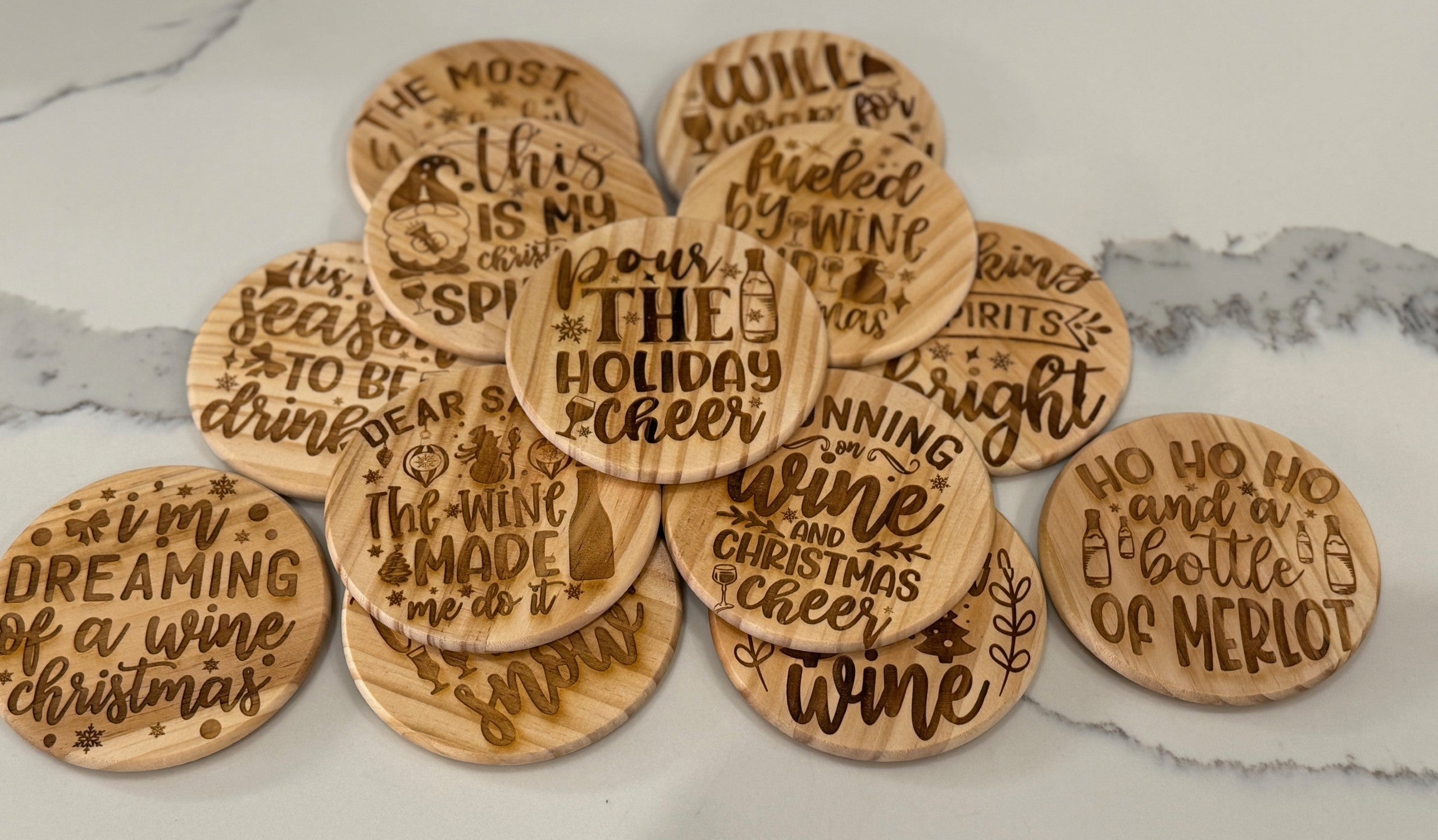 The Funny Wine Sayings: Set of 4 Round Wooden Coasters are arranged on a marble surface. Each coaster features unique festive text like Pour the holiday cheer and Ho ho ho and a bottle of Merlot, adorned with small decorative icons such as stars and wine glasses.