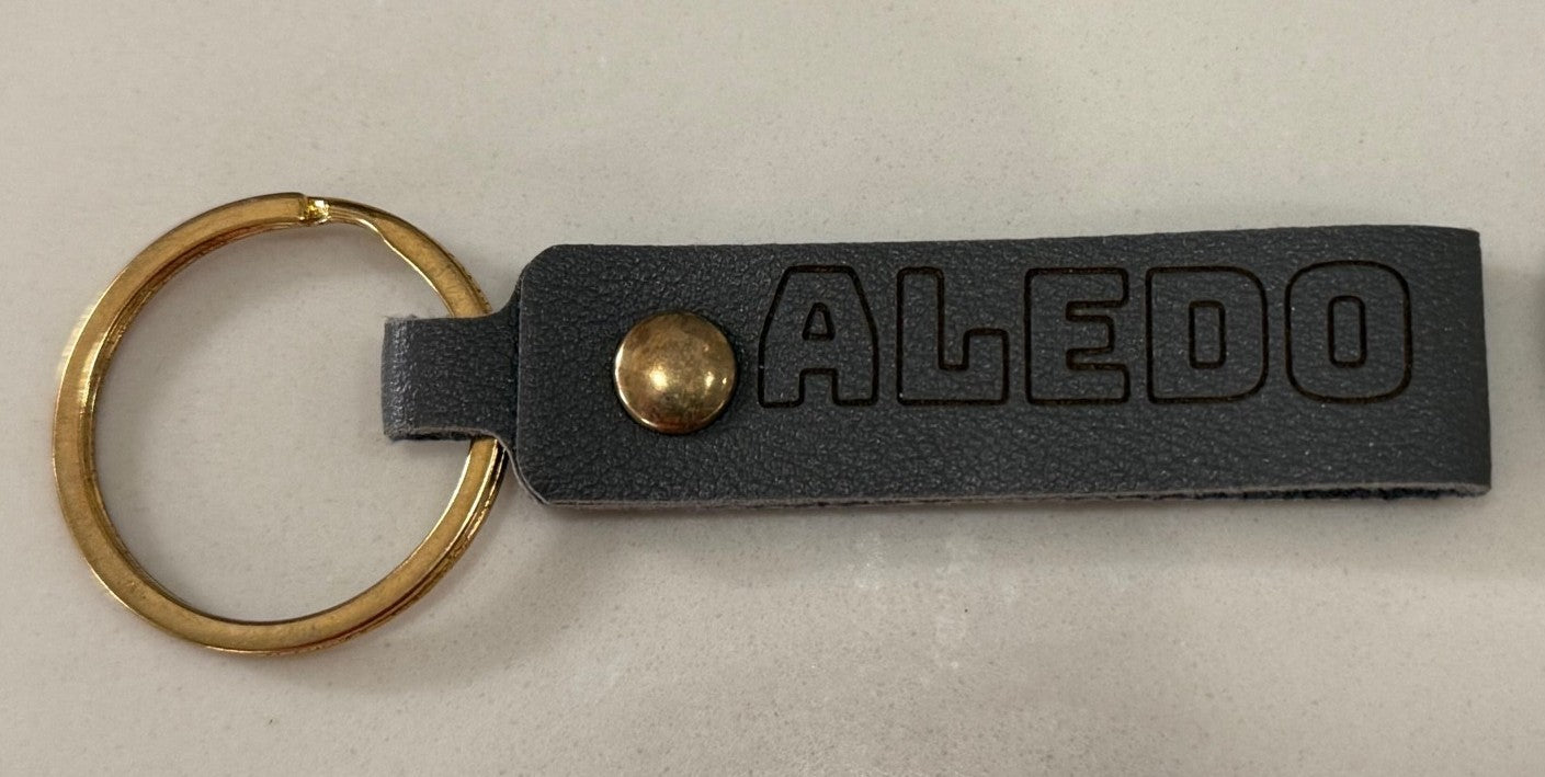 A black leather keychain with the word ALEDO embossed on it in bold letters. The keychain has a brass button and is attached to a gold-colored metal ring. It is lying on a light gray surface.
