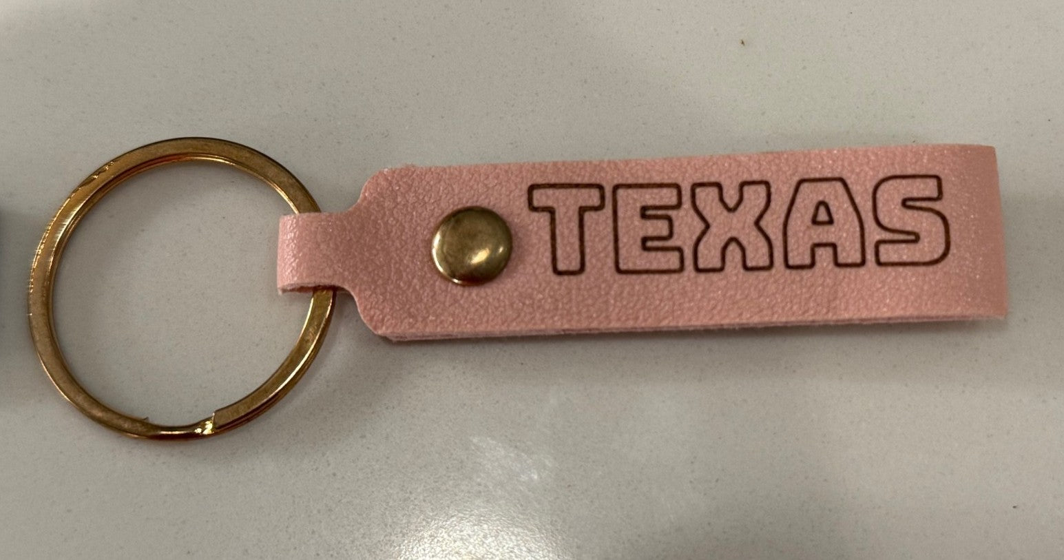 A keychain with a pink leather tag attached to a brass ring. The tag is embossed with the word TEXAS in bold, capital letters. A brass rivet secures the tag to the ring. The keychain is placed on a smooth, light-colored surface.