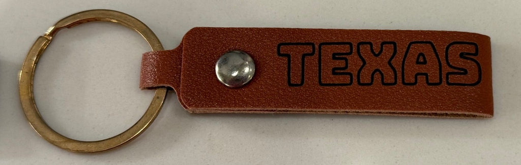 A brown leather keychain with the word TEXAS engraved in bold black letters. It features a shiny metallic snap button and is attached to a gold-colored metal ring. The keychain is laid on a smooth, light-colored surface.