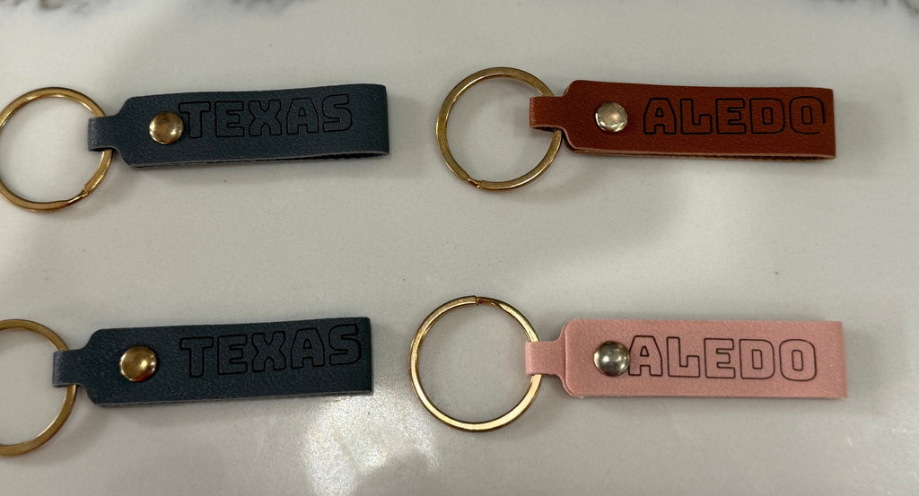 Four leather keychains are displayed on a light surface. Two are dark blue with TEXAS embossed, and two are brown and pink with ALED embossed. Each keychain has a brass ring attached.