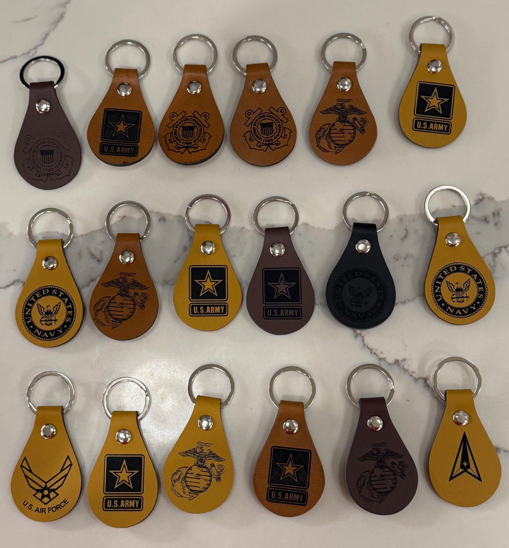 A collection of fifteen teardrop-shaped leather keychains, each embossed with logos from different U.S. military branches, including the Army, Navy, Coast Guard, and Air Force. The keychains vary in color, including brown, tan, yellow, and black, all with metal key rings.