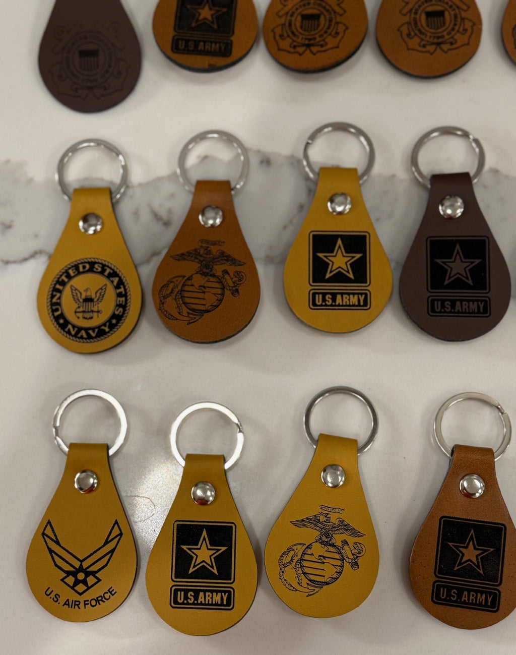 A collection of keychains on a marble surface, featuring logos of U.S. military branches. The keychains are teardrop-shaped in brown and mustard yellow, displaying insignias for the Navy, Marines, Army, and Air Force, each with a silver keyring attached.
