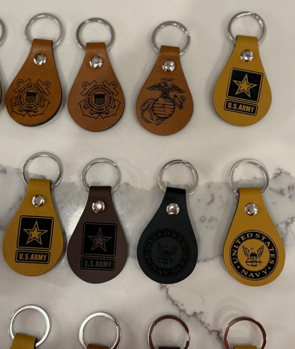 A collection of leather keychains displayed on a marble surface. They feature emblems and logos from various branches of the U.S. military, including the Navy, Marine Corps, and Army. The keychains are in shades of brown, black, and tan, each with a metal ring at the top.