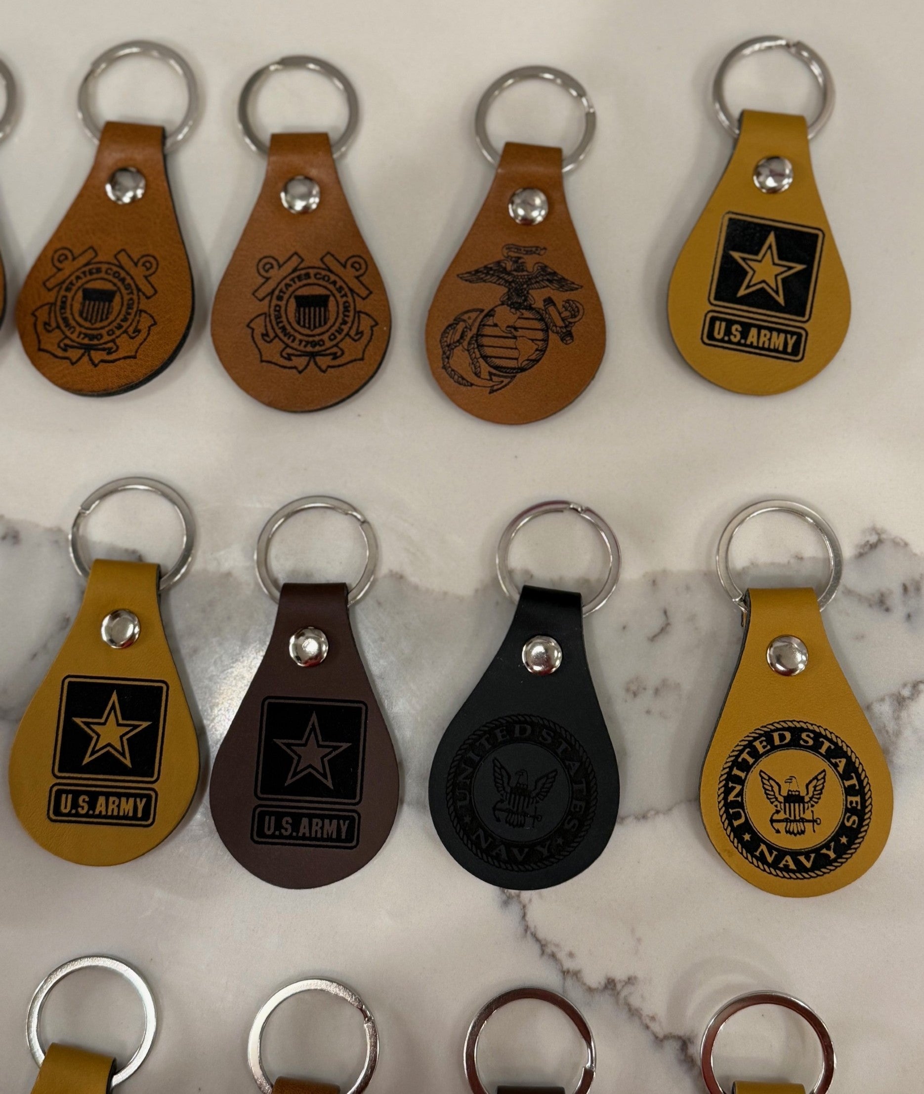 A collection of leather keychains displayed on a marble surface. They feature emblems and logos from various branches of the U.S. military, including the Navy, Marine Corps, and Army. The keychains are in shades of brown, black, and tan, each with a metal ring at the top.