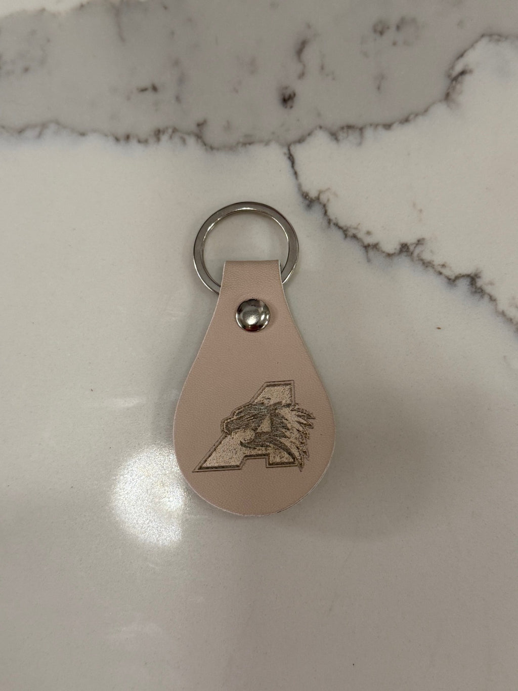 A leather keychain with a teardrop shape rests on a marble surface. It features an engraved letter A with an animal head profile inside it. The keychain is attached to a silver metal ring.