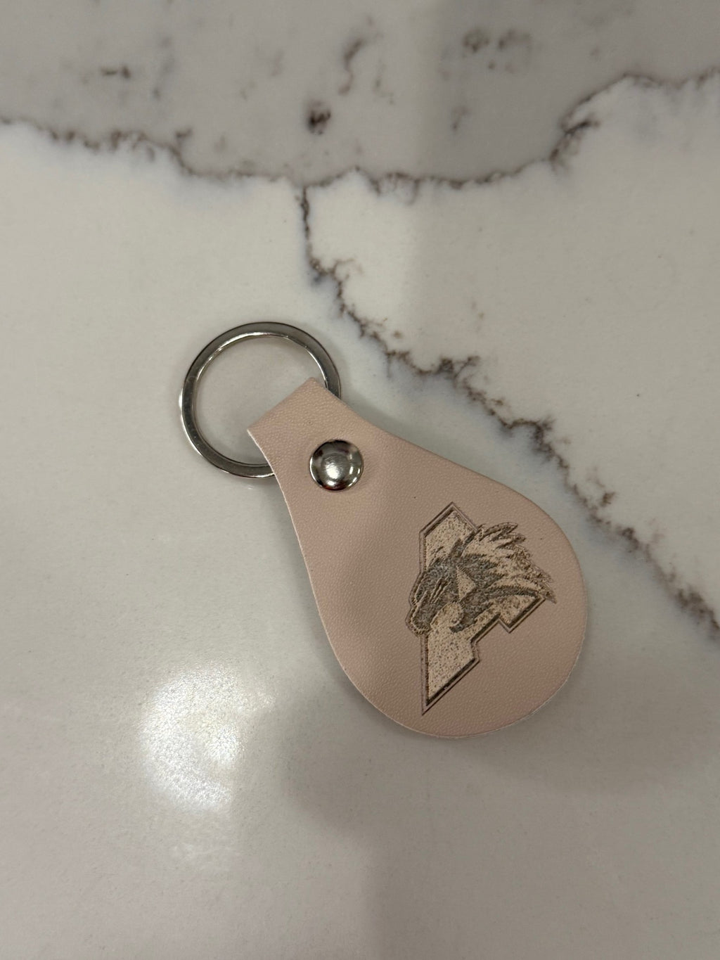A leather keychain on a marble surface. The keychain is beige and shaped like a teardrop, featuring an engraved design of a stylized letter L with an eagle or bird in motion. A metal keyring is attached at the top with a shiny metal rivet.