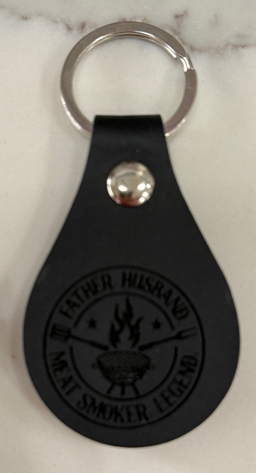 A black leather teardrop-shaped keychain with a silver metal ring on a light surface. The front features an embossed design of a grill with flames and text around it that reads, Father Husband Meat Smoker Legend.