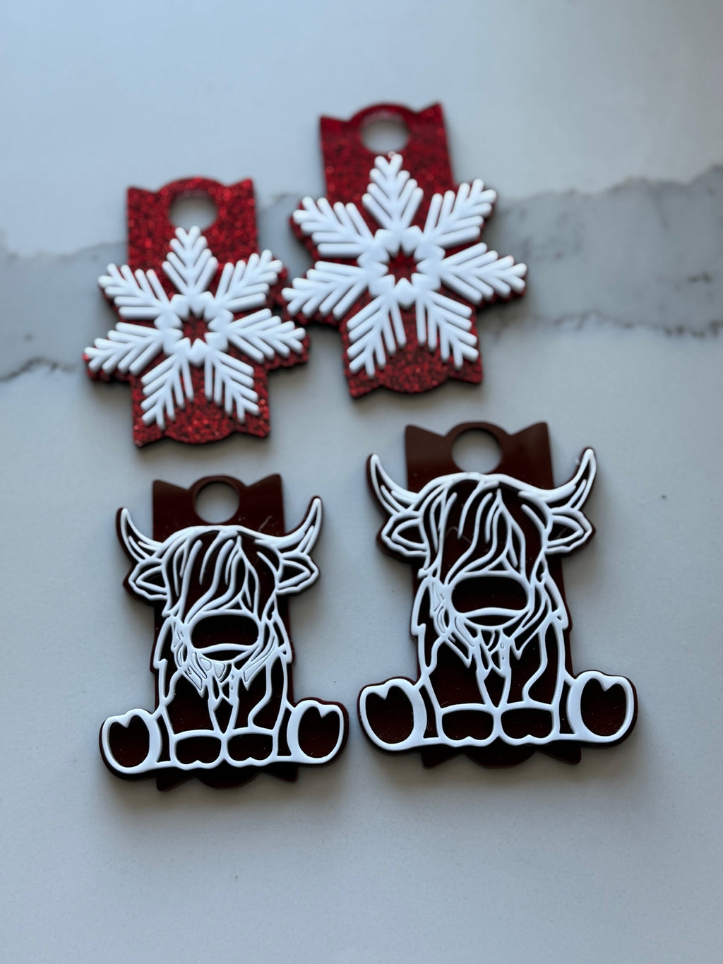 Two pairs of decorative tags sit on a surface. The top pair features red glittery tags with white snowflake designs. The bottom pair shows brown tags with white outlined cartoon cow faces, including horns and a tuft of hair, giving a festive appearance.
